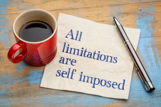 A red cup half-filled with coffee sits on a napkin with the words "All limitations are self-imposed". A pen is opposite the red cup. The cup, the napkin, and the pen are placed on a wood table that has some blue paint on it. 