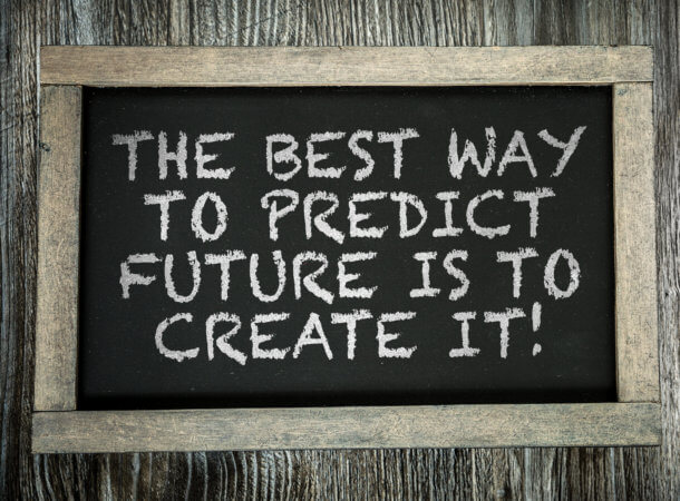 Chalk board with simple wood frame on a wood background.  The comment, in white chalk, is “The best way to predict the future is to create it!”  The picture is in shades of black, grey, and white.