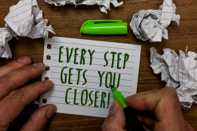 On the top and upper sides of the image are crumpled sheets of paper. In the center, there is a sheet of paper with hands on either side. There is a sharpie in the right hand. On the paper are the words “Every Step Gets You Closer”. 