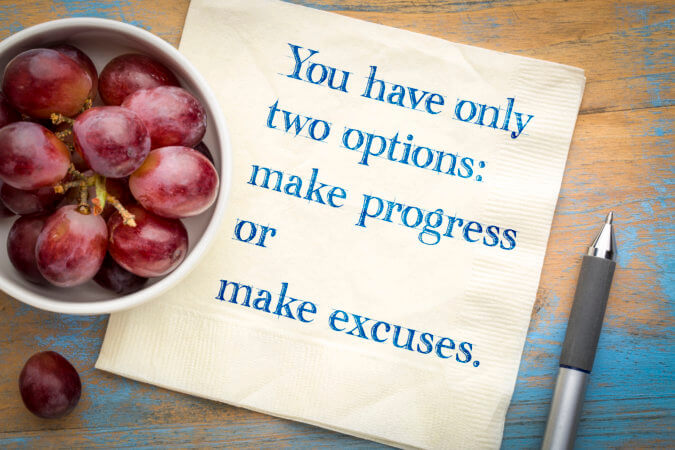 A bowl of cherries is on the left side. One cherry is out of the bowl and is below the bowl, on the table. To the right of the bowl is a dinner napkin. On the napkin the following words are printed in blue: “You have only two options; make progress or make excuses.” On the right of the napkin is a ball point pen. 