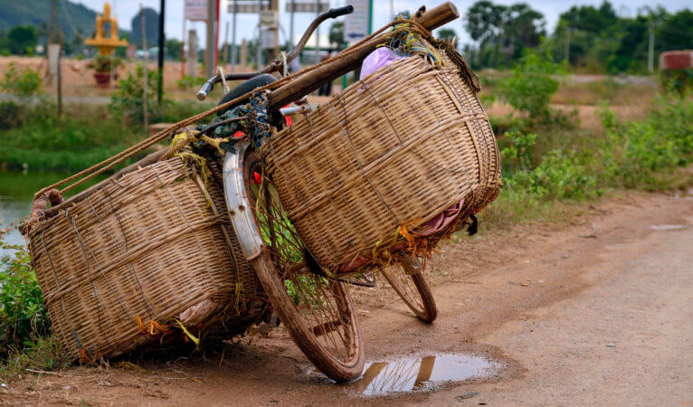 A bicycle has two baskets, on one each side, hanging from a pole behind the bicycle seat. The baskets are full. The bike has no rider and is tilted, leaning on a basket on a dirt road. There is a puddle of water under the back wheel. There is a pond to the left, and equipment of some sort in the distant background. 