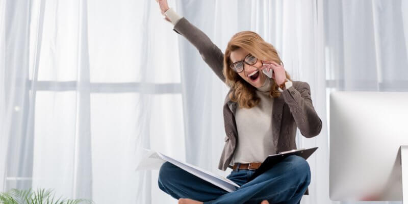 Woman who is wearing jeans, a white shirt and grey jacket is sitting cross-legged on a table/desk. Her right arm is raised and she is talking on the phone. She appears happy.