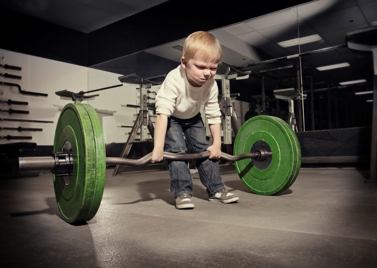 A young blond boy, wearing jeans and a long-sleeved tee shirt, is in a gym. He is trying to lift a weight that weighs more than he does. He has a determined look on his face. 