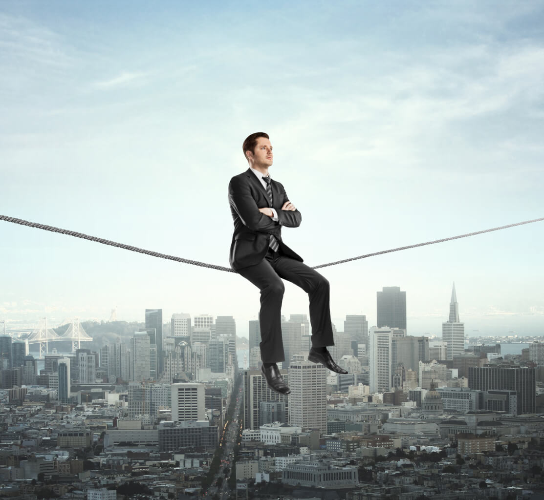 There is a picture of a metropolitan city skyline in the background.In the foreground, a man in a suit, with his arms crossed in front of him, is sitting on a tightrope. He seems perfectly calm and self-assured. 