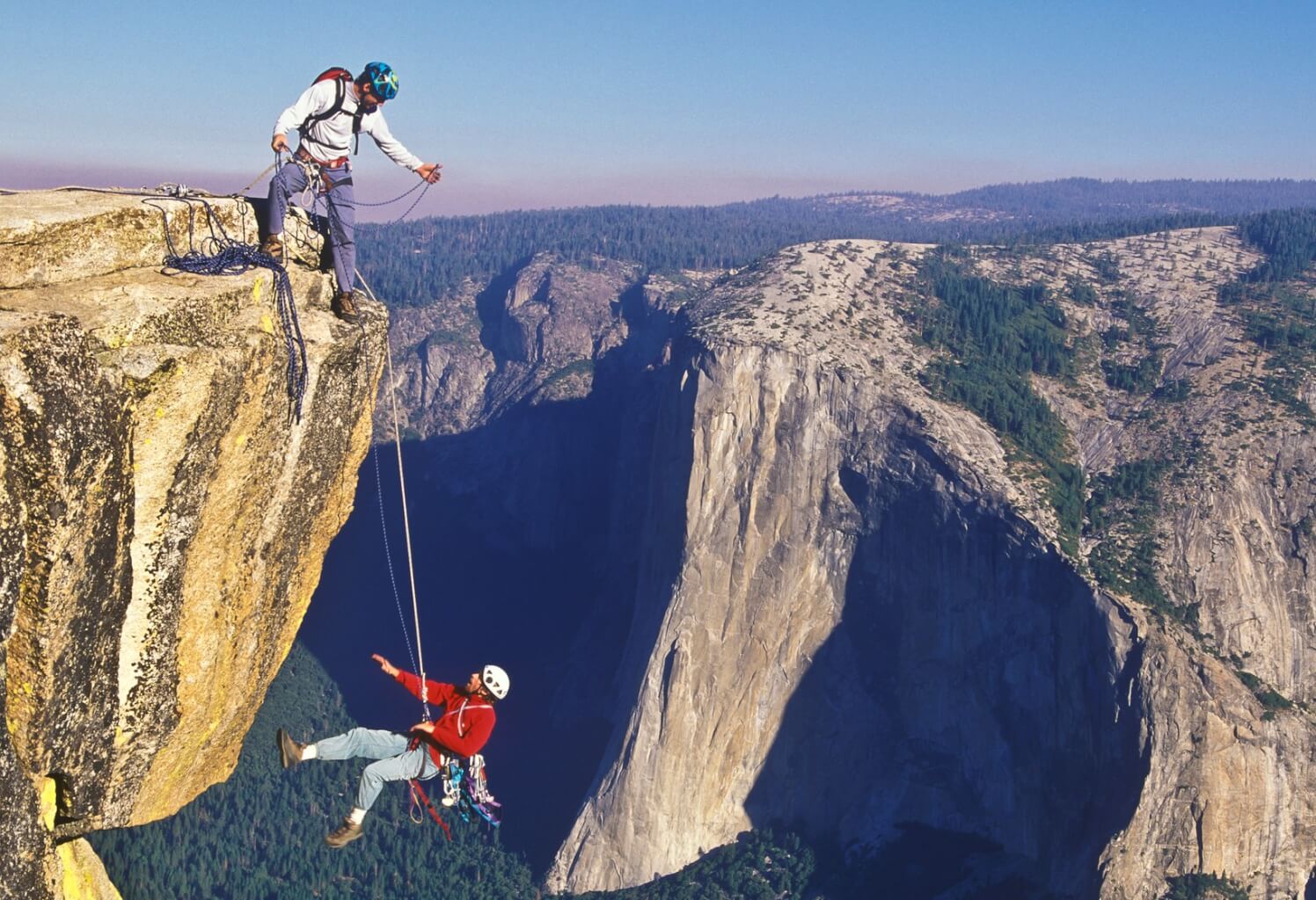 Mountainous area with two people climbing a rock. One person is standing on a rock formation, and another is on a cord, dangling below the person standing on the rock formation.