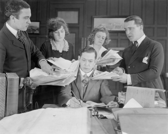 Man in a suit sitting at a table has four people behind him. A man is on the far left and the far right sides. Two women are in the middle. Each of these four people are shoving papers toward the man who is trying to work on something on the table in front of him. 