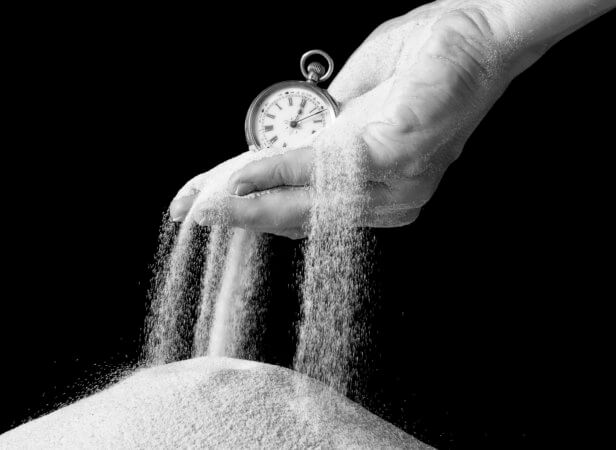 This is a black and white photo. There is a hand holding a pocket watch and sand is spilling out of the hand onto a pile of sand below the hand. This is to convey the idea that time slips away.