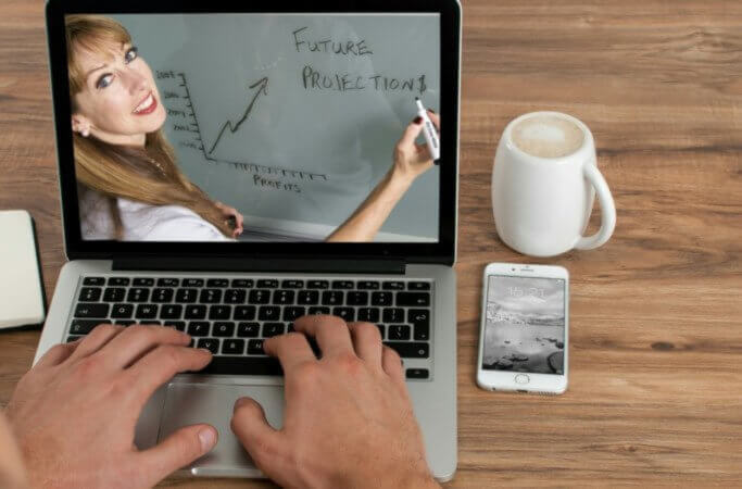 Laptop with hands on keyboard; picture of woman on the screen writing "future projections"; cup with beverage and phone next to laptop on wood grain surface
