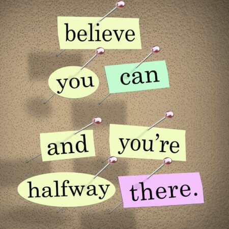 Grey to black background. The words “Believe you can and you’re halfway there” are on the graphic. Each word is on a lighter color, as if written on a post-it note. The words are, however, “attached” to the grey/black background with pins. 