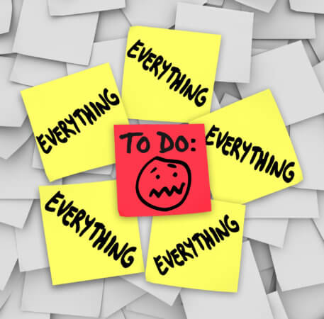 The background is a collection of miscellaneously placed white squares that look like sticky notes. On top of that pile are five yellow sticky notes with the word “Everything” hand printed on each note. In the center, there is one red square with a cartoon face in the middle and the words “To Do” above the face. The face seems overwhelmed. 