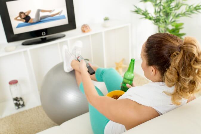Woman on couch, with soda in her right hand and television remote in her left hand. Her legs are crossed and her feet are resting on an exercise ball. She is watching an exercise video on a screen in front of her. 