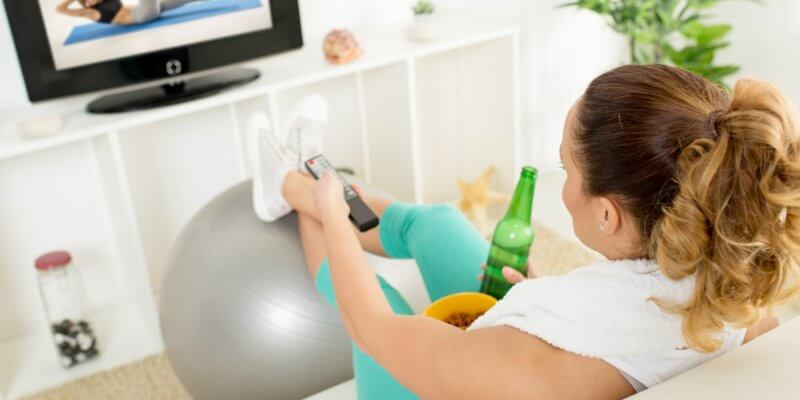 Woman on couch, with soda in her right hand and television remote in her left hand. Her legs are crossed, and her feet are resting on an exercise ball. She is watching an exercise video on a screen in front of her.