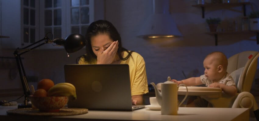 Medium shot of a young mother working on a laptop while her baby is sitting next to her. It is night time and she appears to be at a kitchen table since there is a bowl of fruit in front of her laptop. She has her right hand covering the bottom half of her face. There is what appears to be a single light bulb lighting the woman’s laptop. 