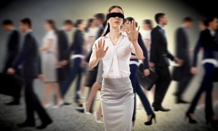 Fuzzy, blurred group of people walking left and right in the background. In the foreground, a young woman in white blouse and grey skirt is blindfolded and feeling her way toward the viewer/reader.